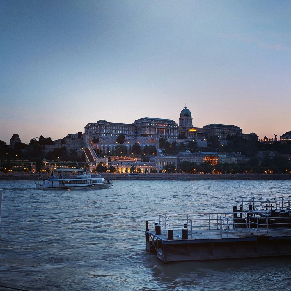Der Budapester Burgpalast über der Donau in der Dämmerung, davor ein Ausflugsschiff auf dem Wasser