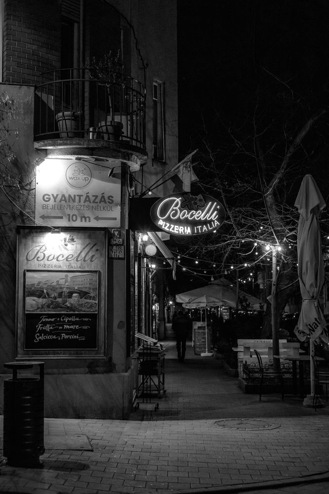 Straßenecke in Budapest bei Nacht in Schwarzweiß, Leuchtschrift einer Pizzeria, Lichterketten und Gäste im Hintergrund