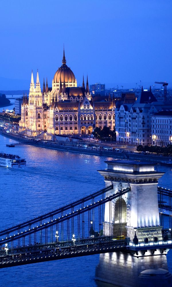 Blick über die Donau auf das beleuchtete ungarische Parlament, im Vordergrund der Pfeiler der Kettenbrücke zur blauen Stunde